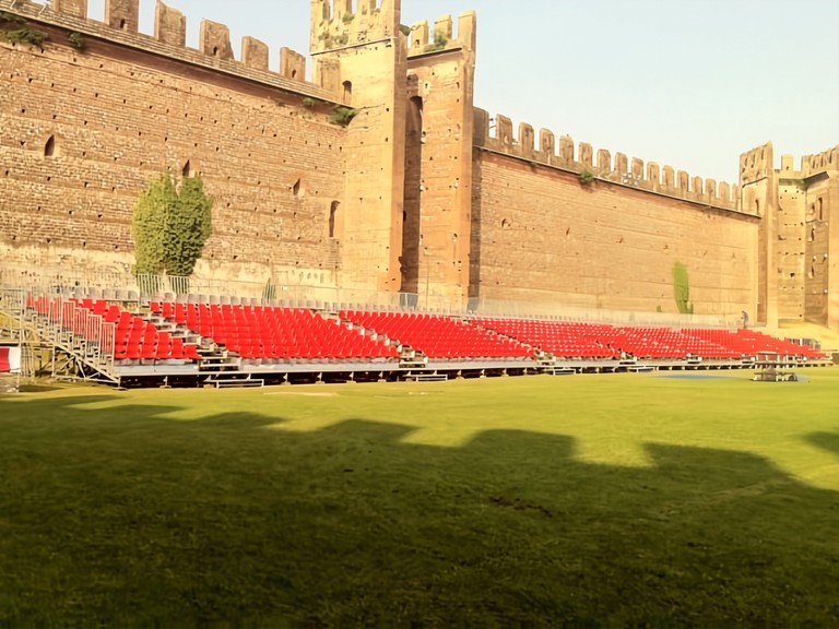 Tribune con sedute rosse montate su un prato ai piedi di antiche mura merlate